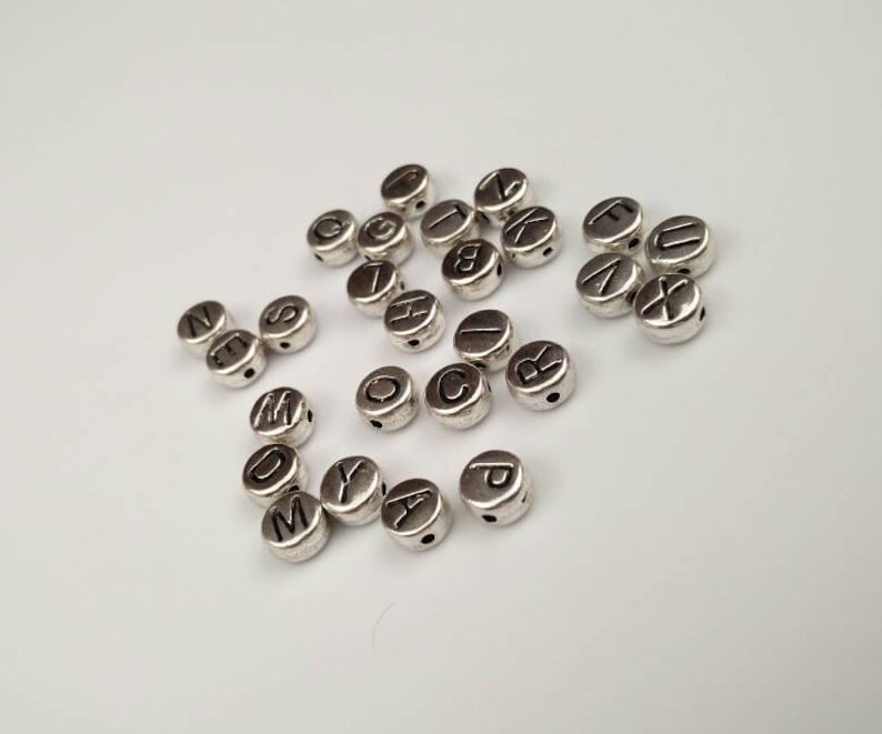 May include: Silver letter beads with black lettering. The beads are round and have a hole in the center. The letters are A through Z.