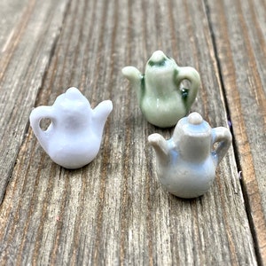 May include: Three miniature ceramic teapots in pastel shades: light blue, pale green, and a blue-grey. Each teapot has a small spout, handle, and lid. The teapots are arranged on a weathered, wooden surface.
