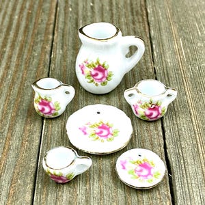 Puede incluir: Un juego de té en miniatura con fondo blanco y diseño floral de rosas rosas. El juego incluye una jarra, dos tazas, dos platillos y un plato.