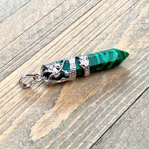 Op de afbeelding: Een groene en zwarte malachiet kristallen hanger met een zilverkleurig drakenontwerp. Het kristal heeft een gepolijste, langwerpige vorm met een puntig uiteinde. De hanger heeft een zilverkleurige beugel om aan een ketting te bevestigen.