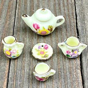 May include: A miniature floral tea set with a teapot, creamer, sugar bowl, cup and saucer. The set is white with gold trim and features a floral pattern in shades of pink, yellow, and purple.