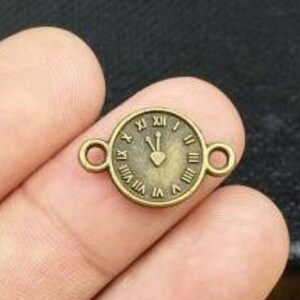 Puede incluir: Un dije de reloj de metal de color bronce con números romanos y un diseño de corazón. El dije tiene dos anillos de salto para sujetarlo a joyas u otras manualidades.