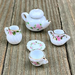 Puede incluir: Un juego de té en miniatura con una tetera blanca, jarra de crema, azucarero, platillo y taza de té. El juego está decorado con flores rosas y hojas verdes.