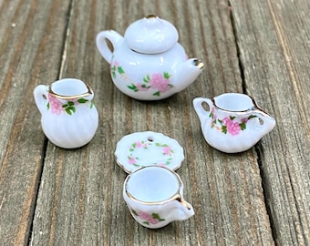 May include: A miniature tea set with a white teapot, creamer, sugar bowl, saucer, and teacup. The set is decorated with pink flowers and green leaves.