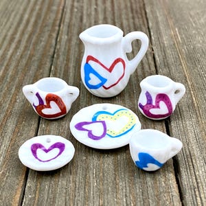 Puede incluir: Un juego de té en miniatura con una tetera blanca, dos tazas, dos platillos y un plato. El juego está decorado con corazones pintados a mano en varios colores, incluyendo rojo, azul, morado y amarillo.