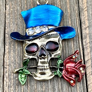 May include: A gold-toned skull charm with a blue top hat adorned with rhinestones. The skull is holding a red rose in its mouth.