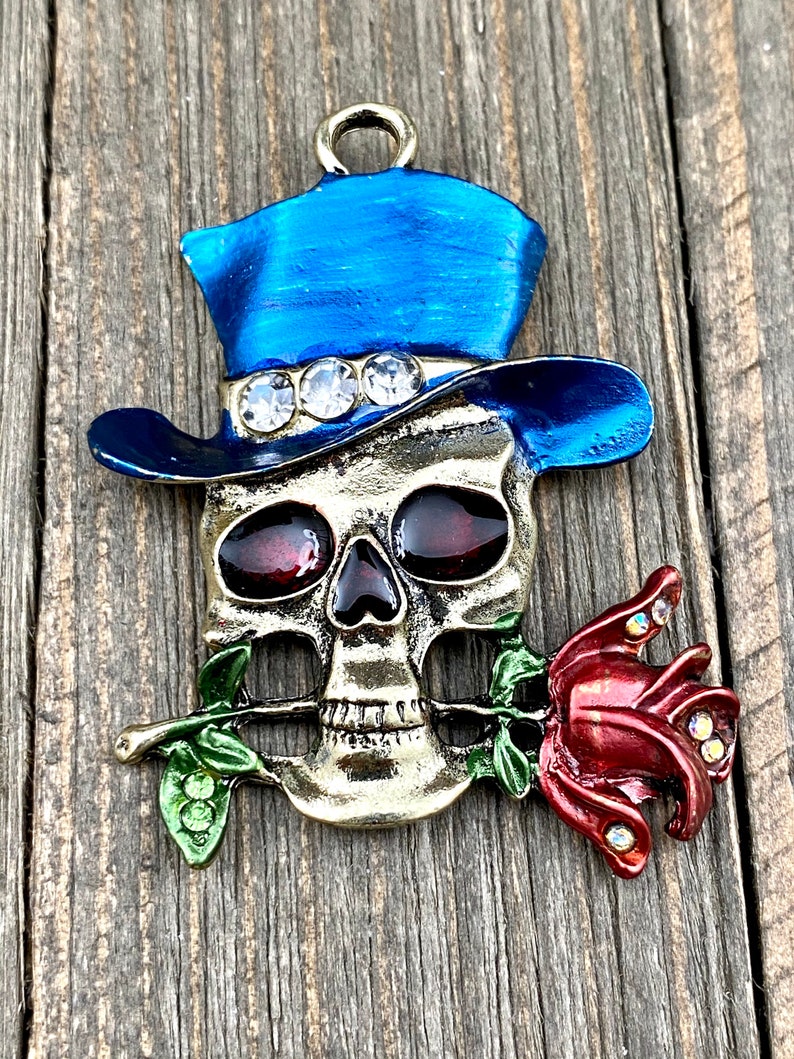 May include: A gold-toned skull charm with a blue top hat adorned with rhinestones. The skull is holding a red rose in its mouth.