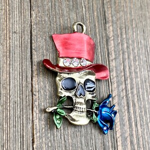 May include: A gold-toned skull charm with a red top hat and a blue rose in its mouth. The hat has rhinestones around the band. The skull has black eyes and a white smile.