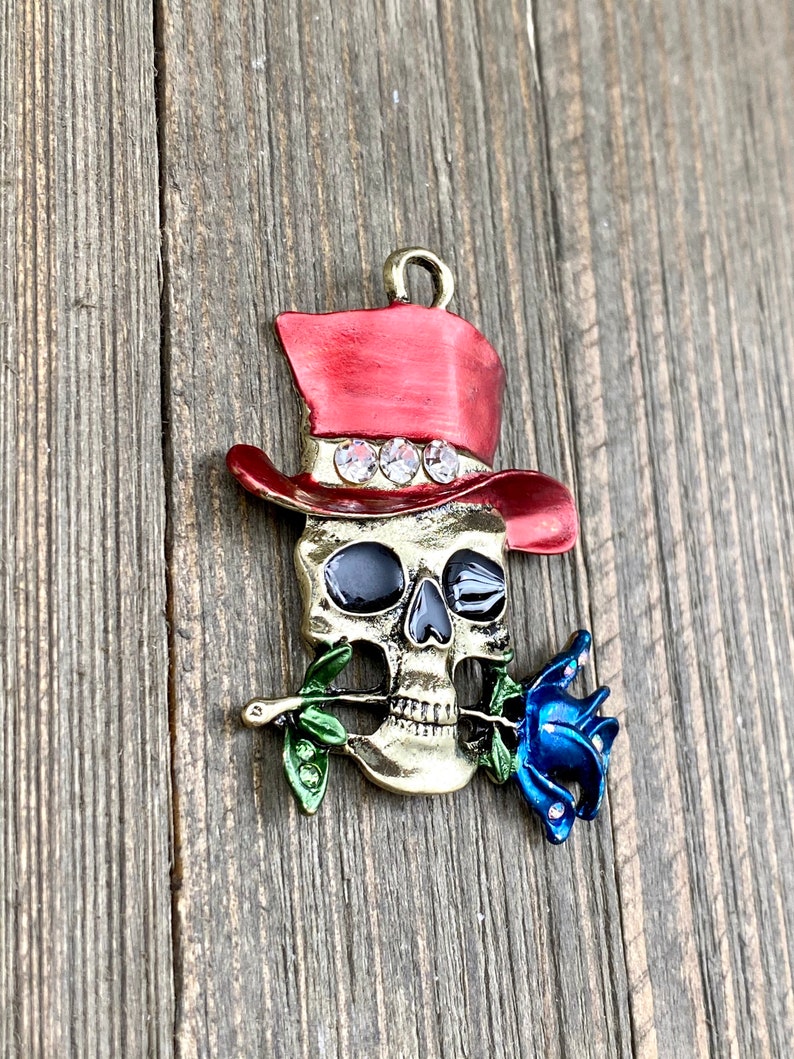 May include: A gold-toned skull charm with a red top hat and a blue rose in its mouth. The hat has rhinestones around the band. The skull has black eyes and a white smile.