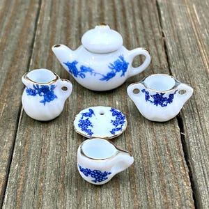 Puede incluir: Un juego de té de porcelana en miniatura con diseños florales azules y dorados. El juego incluye una tetera, una jarra de crema, un azucarero, un platillo y una taza de té.