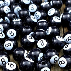 May include: A collection of black and white eight ball beads. Each bead features a glossy black finish with a white circle displaying the number eight. The beads are round and have a hole for stringing.