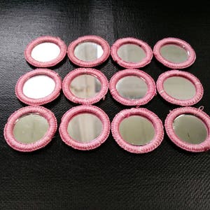 Puede incluir: Doce pequeños espejos redondos con marcos rosas tejidos sobre una superficie negra. Los espejos reflejan el entorno. Los marcos rosas contrastan con el fondo oscuro, añadiendo un elemento decorativo.