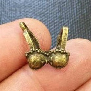 Könnte beinhalten: Ein goldfarbener Metallanhänger in Form eines BHs. Der Anhänger hat ein detailliertes Design mit einer Perlenbordüre.