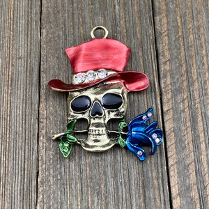 May include: A gold-toned skull charm with a red top hat and a blue rose. The skull has black eyes and a white trim around the hat.