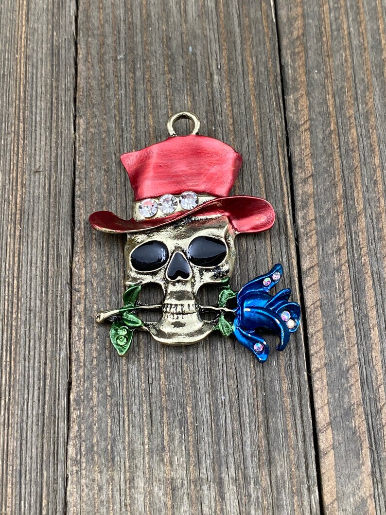 May include: A gold-toned skull charm with a red top hat and a blue rose. The skull has black eyes and a white trim around the hat.
