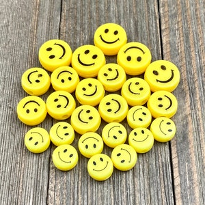 Puede incluir: Una colección de cuentas amarillas con caritas sonrientes y detalles negros. Las cuentas están dispuestas en forma de corazón sobre una superficie de madera.