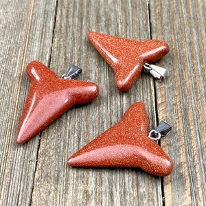 May include: Three red sandstone shark tooth pendants with silver bail. The pendants are on a wooden surface.