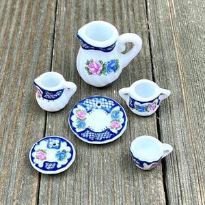 Puede incluir: Un juego de té en miniatura con base blanca y diseños florales azules y rosas. El juego incluye una tetera, una jarra de crema, un azucarero y dos tazas de té y platillos.