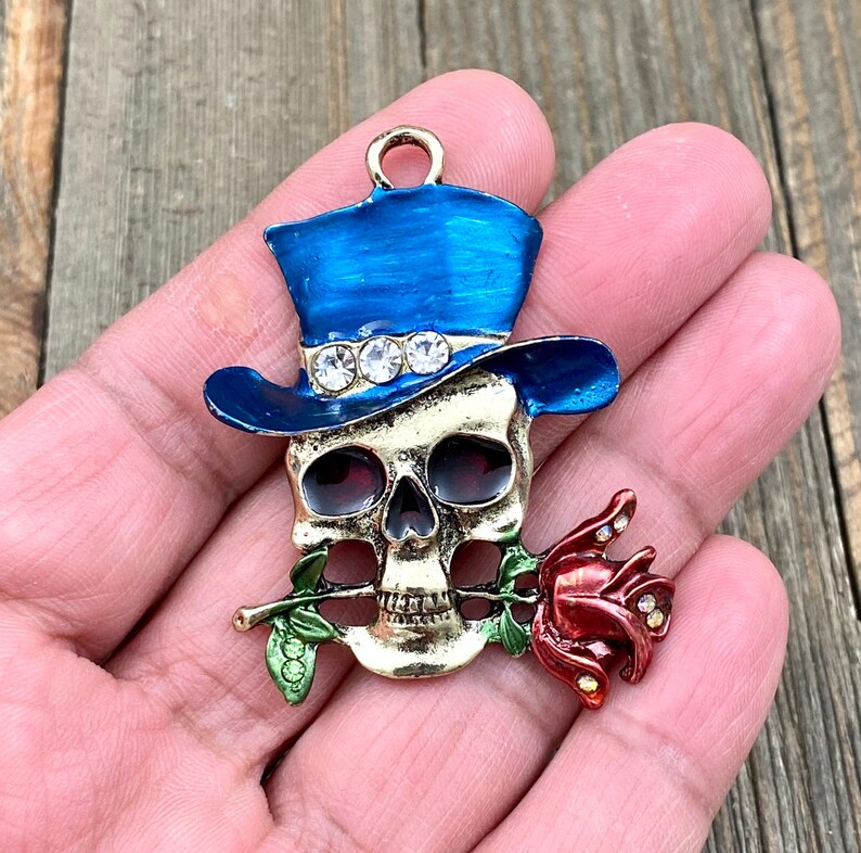 May include: A gold-toned skull charm with a blue top hat and a red rose. The hat has rhinestones around the band. The skull is holding the rose in its mouth.