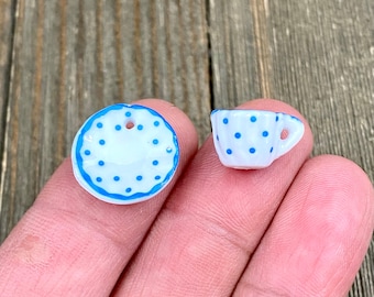 Puede incluir: Miniatura de taza y platillo blancos con lunares azules. El platillo tiene un borde azul y un agujero para ensartar. La taza tiene un asa. El juego se sostiene en la mano de una persona.