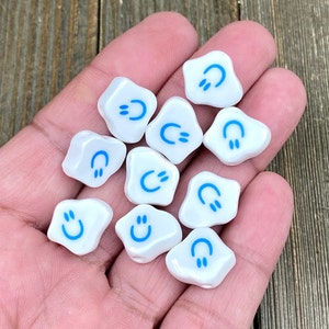 May include: A collection of white, irregularly shaped beads with a glossy finish. Each bead features a blue smiley face design. The beads are small, suitable for jewelry making or craft projects. The background is a wooden surface.