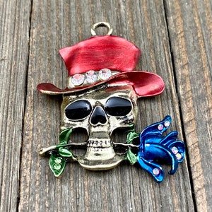 May include: A gold-tone skull charm with a red cowboy hat, black eyes, and a blue rose in its mouth. The hat is decorated with rhinestones.