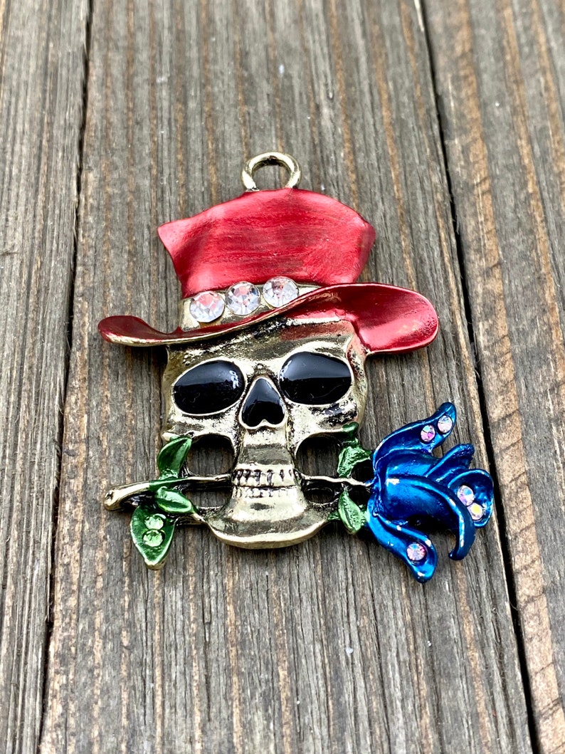 May include: A gold-tone skull charm with a red cowboy hat, black eyes, and a blue rose in its mouth. The hat is decorated with rhinestones.