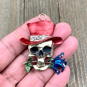 May include: Gold-tone skull charm with a red top hat, blue rose, and green stem. The skull has black eyes and rhinestones around the hat.