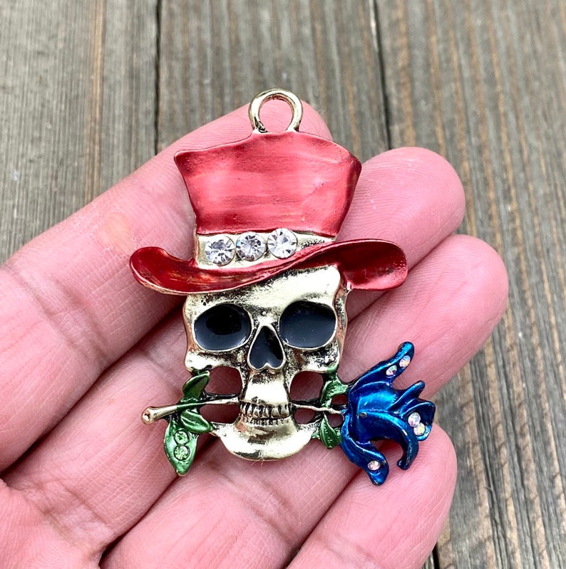 May include: Gold-tone skull charm with a red top hat, blue rose, and green stem. The skull has black eyes and rhinestones around the hat.