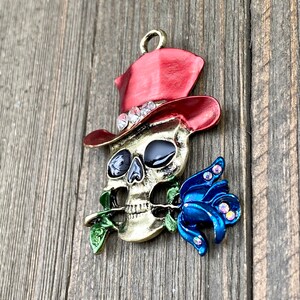 May include: A gold-colored skull wearing a red top hat with rhinestones and holding a blue rose with rhinestones.