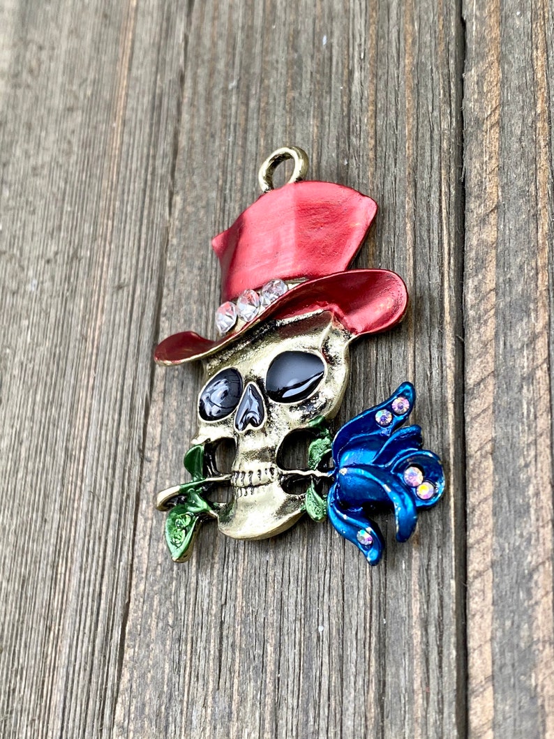 May include: A gold-colored skull wearing a red top hat with rhinestones and holding a blue rose with rhinestones.