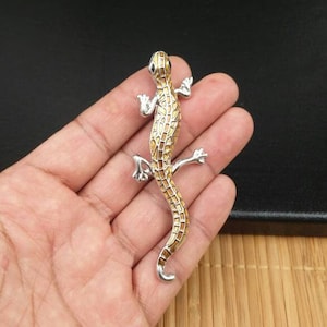 May include: A silver and gold lizard brooch with a detailed, textured design. The lizard is standing on its hind legs with its tail curled up.