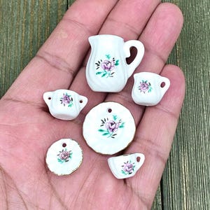 Puede incluir: Un juego de té en miniatura con fondo blanco y diseño floral de rosas rosas. El juego incluye una jarra, dos tazas, dos platillos y un plato.