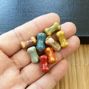 May include: A collection of colorful ceramic bone-shaped beads. The beads are in various shades of blue, green, yellow, orange, and brown. They are perfect for jewelry making or other craft projects.