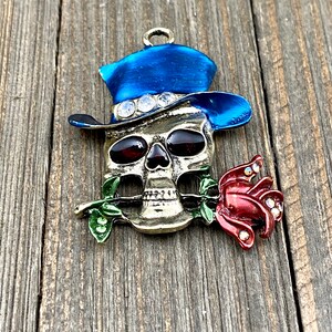 May include: A gold-toned skull charm with a blue top hat and a red rose. The skull has red eyes and the hat has rhinestones.