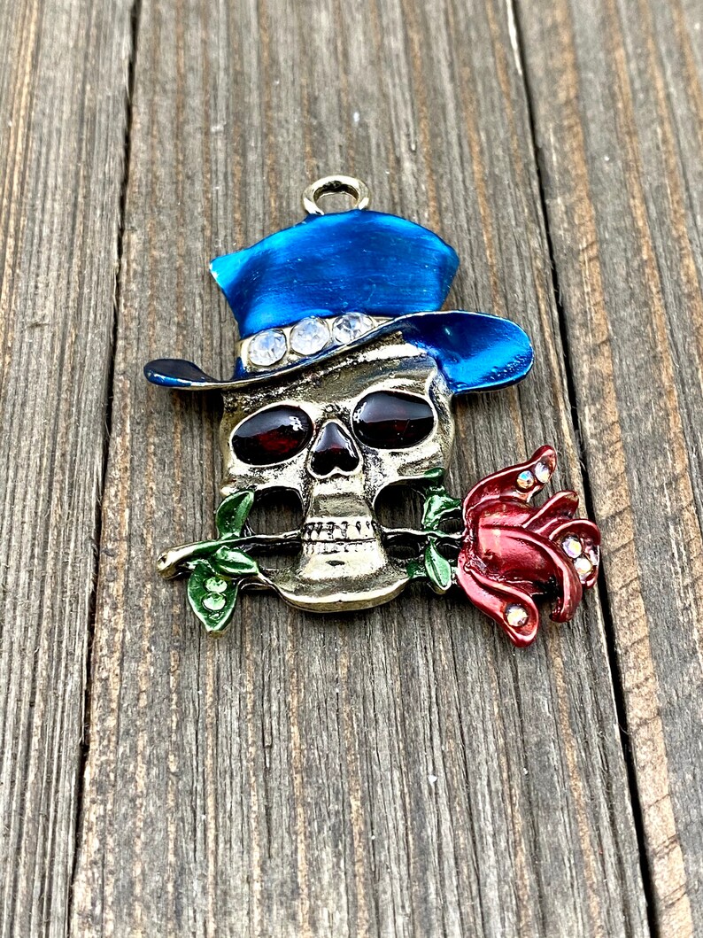 May include: A gold-toned skull charm with a blue top hat and a red rose. The skull has red eyes and the hat has rhinestones.