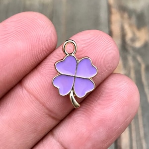 May include: A gold-colored four-leaf clover charm with a purple enamel finish. The charm has a loop at the top for attaching to a necklace or bracelet.