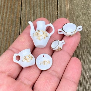 May include: A miniature white porcelain tea set with gold floral detailing. The set comprises a teapot, creamer, sugar bowl, two teacups, and a saucer. The pieces are small, delicate, and appear handmade.