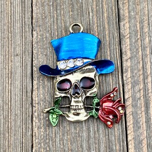 May include: A gold-toned skull charm with a blue top hat and a red rose. The skull has red eyes and the rose has rhinestones.