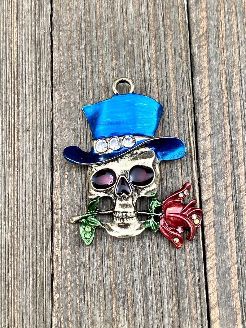 May include: A gold-toned skull charm with a blue top hat and a red rose. The skull has red eyes and the rose has rhinestones.
