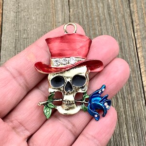 May include: A gold-tone skull charm with a red top hat and a blue rose. The skull has black eyes and a white band around the hat with rhinestones.