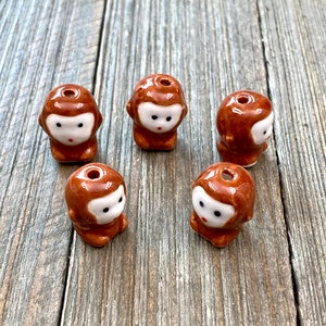 May include: Five brown ceramic monkey beads with white faces and black eyes. The beads have a hole through the top for stringing.