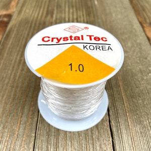 Może przedstawiać: Biała szpula z przezroczystym elastycznym sznurkiem Crystal Tec o grubości 1,0 mm, wyprodukowana w Korei. Szpula jest oznaczona nazwą marki i rozmiarem.