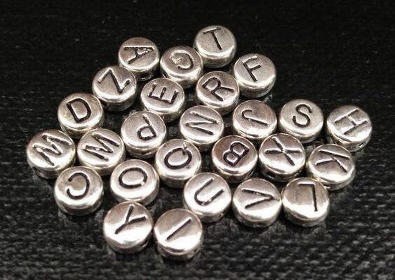 May include: A collection of silver metal alphabet letter beads. The beads are round and have a smooth surface. Each bead has a single letter engraved on it.
