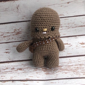 May include: A brown crocheted plush toy resembling a character from Star Wars. The toy has black eyes, a white mouth, and a brown bandolier.
