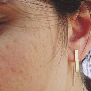 May include: A person wearing a pair of gold bar earrings. The earrings are long and thin, and they hang down from the earlobes.