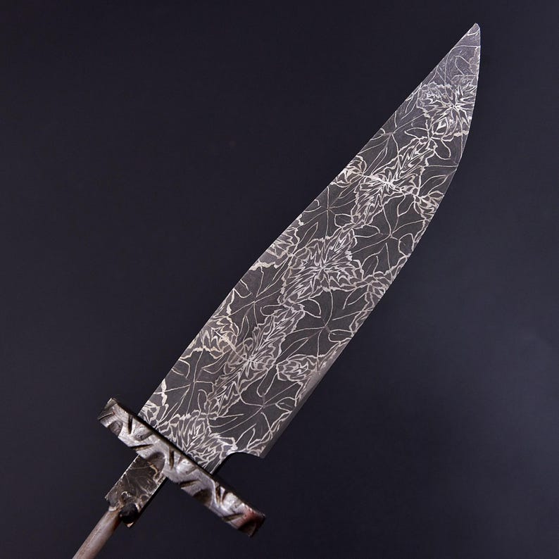 May include: A steel blade with a floral pattern etched across its surface. The blade has a long, pointed shape with a cross-shaped handle. The floral design is intricate and detailed, creating a decorative effect. The blade is set against a dark background.