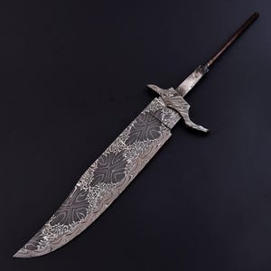 Può includere: Una grande lama di coltello incompiuta con un motivo decorativo inciso nell'acciaio. La lama ha un profilo ampio e una punta. Il codolo si estende in un'asta filettata stretta. Il metallo ha un aspetto scuro e strutturato.