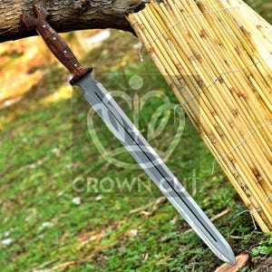 May include: A long, silver sword with a wooden handle featuring intricate carvings. The sword is resting on a bed of green grass and a woven bamboo mat.