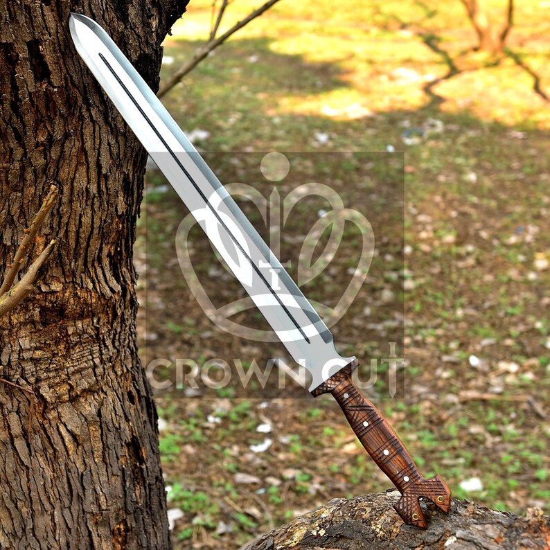 May include: A long, silver sword with a wooden handle. The handle is decorated with a brown and black pattern. The sword is resting on a tree trunk.